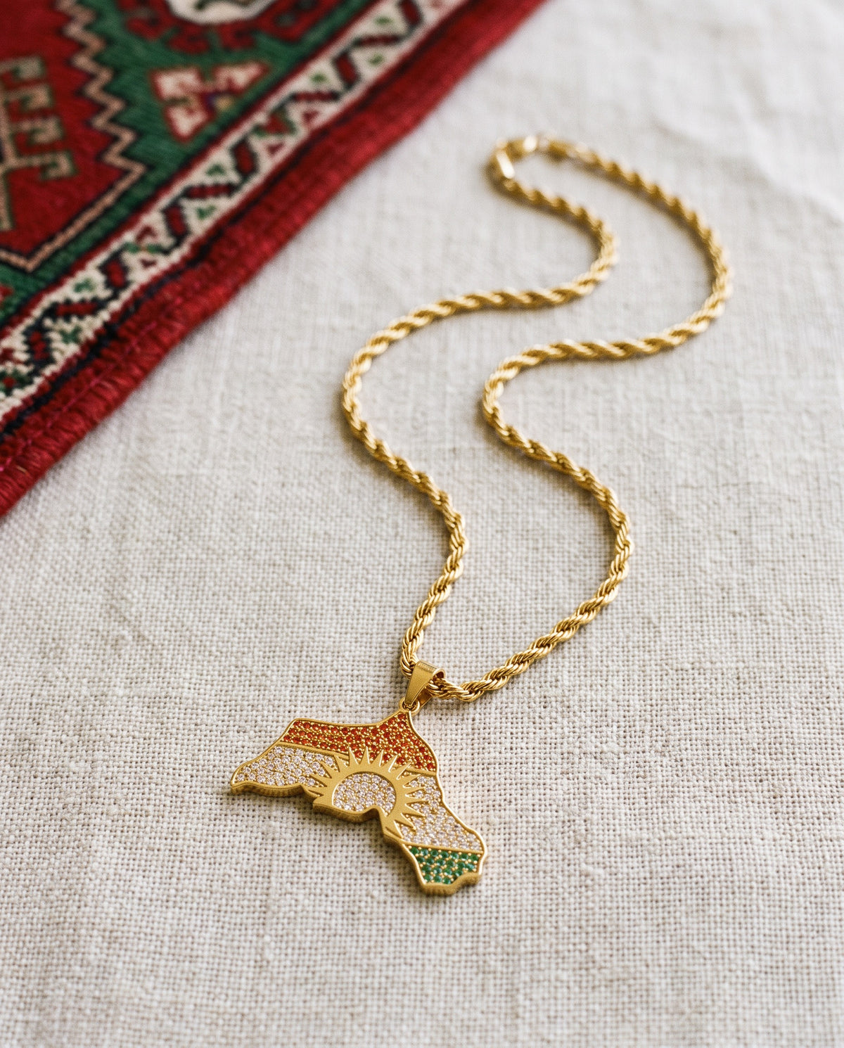 Kurdistan necklace for men in 18K gold, map of Kurdistan pendant with red, white and green zirconia around the sun, rope chain, flat lay on linen with Kurdish textile