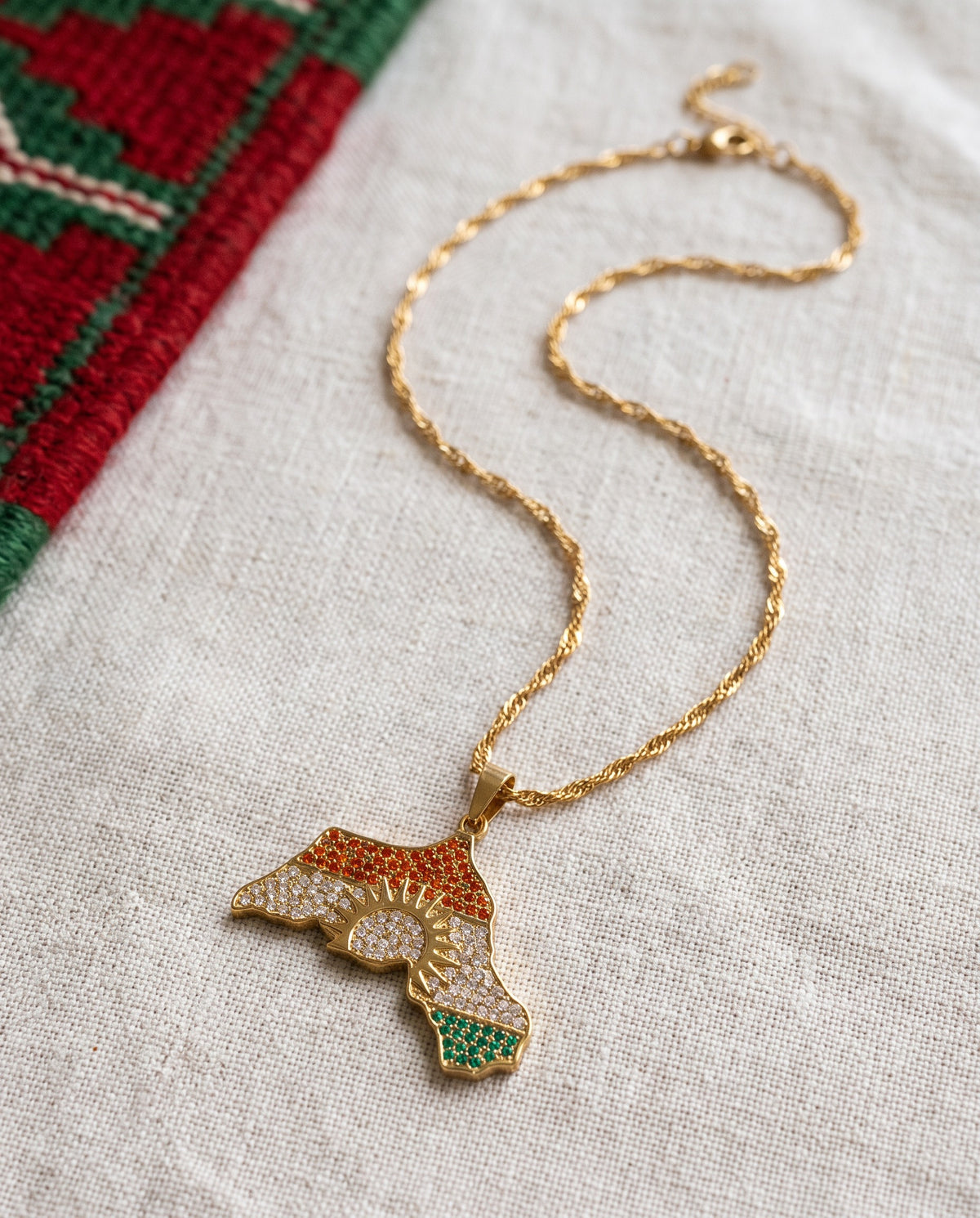 Kurdistan necklace for women in 18K gold, map of Kurdistan pendant with red, white and green zirconia around the sun, fine wave chain, flat lay on linen with Kurdish textile