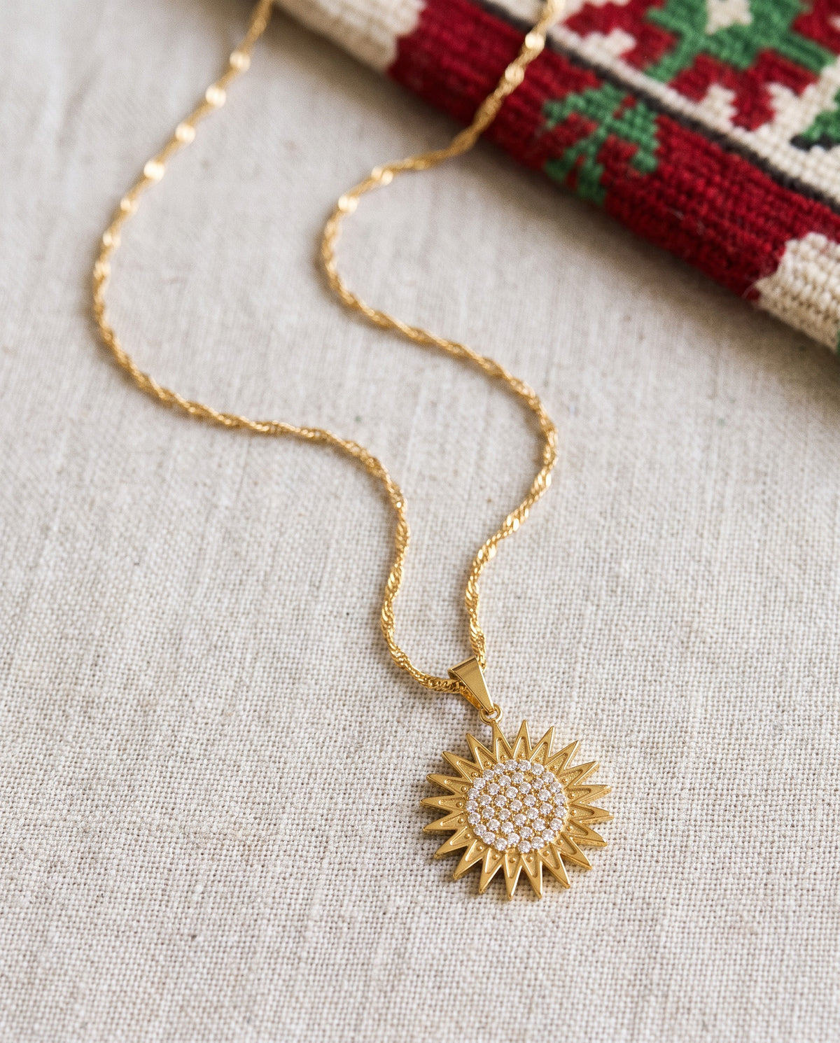 Kurdistan necklace for women in 18K gold, Kurdish sun pendant with 21 rays and clear zirconia, fine wave chain, flat lay on linen with Kurdish textile