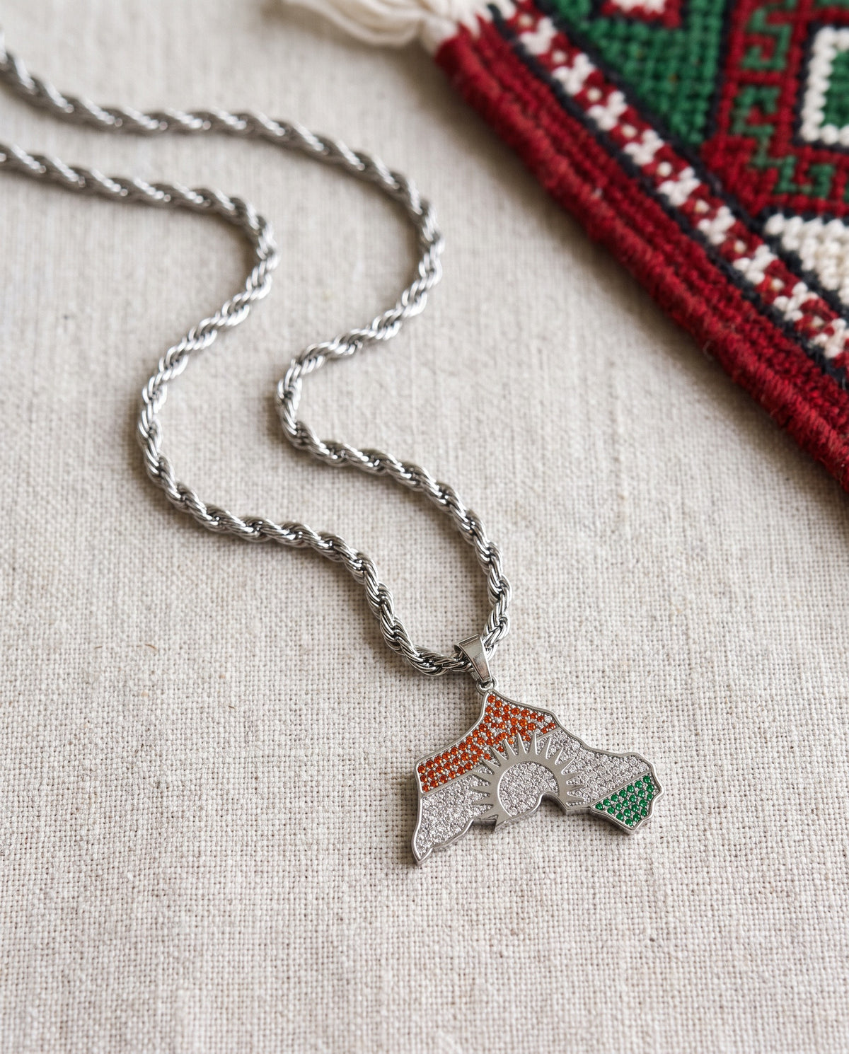 Kurdistan necklace for men in silver, map of Kurdistan pendant with red, white and green zirconia around the sun, rope chain, flat lay on linen with Kurdish textile