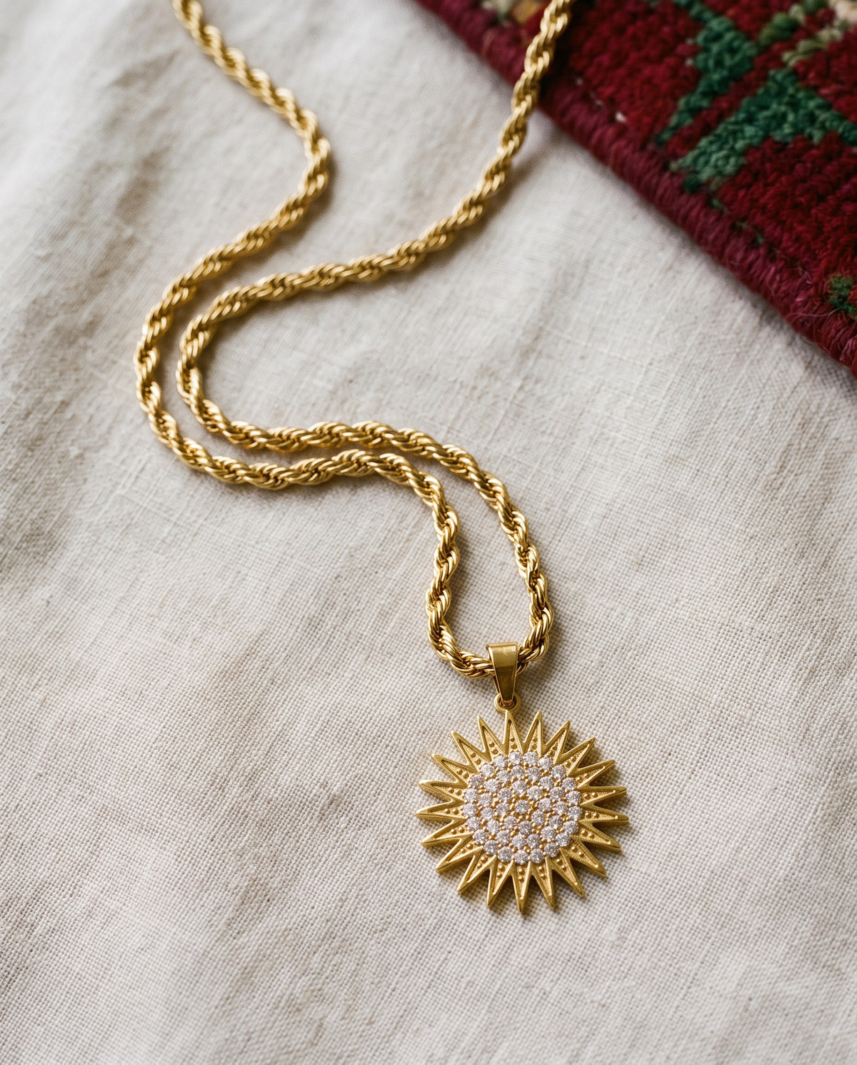 Kurdistan necklace for men in 18K gold, Kurdish sun pendant with 21 rays and clear zirconia, rope chain, flat lay on linen with Kurdish textile